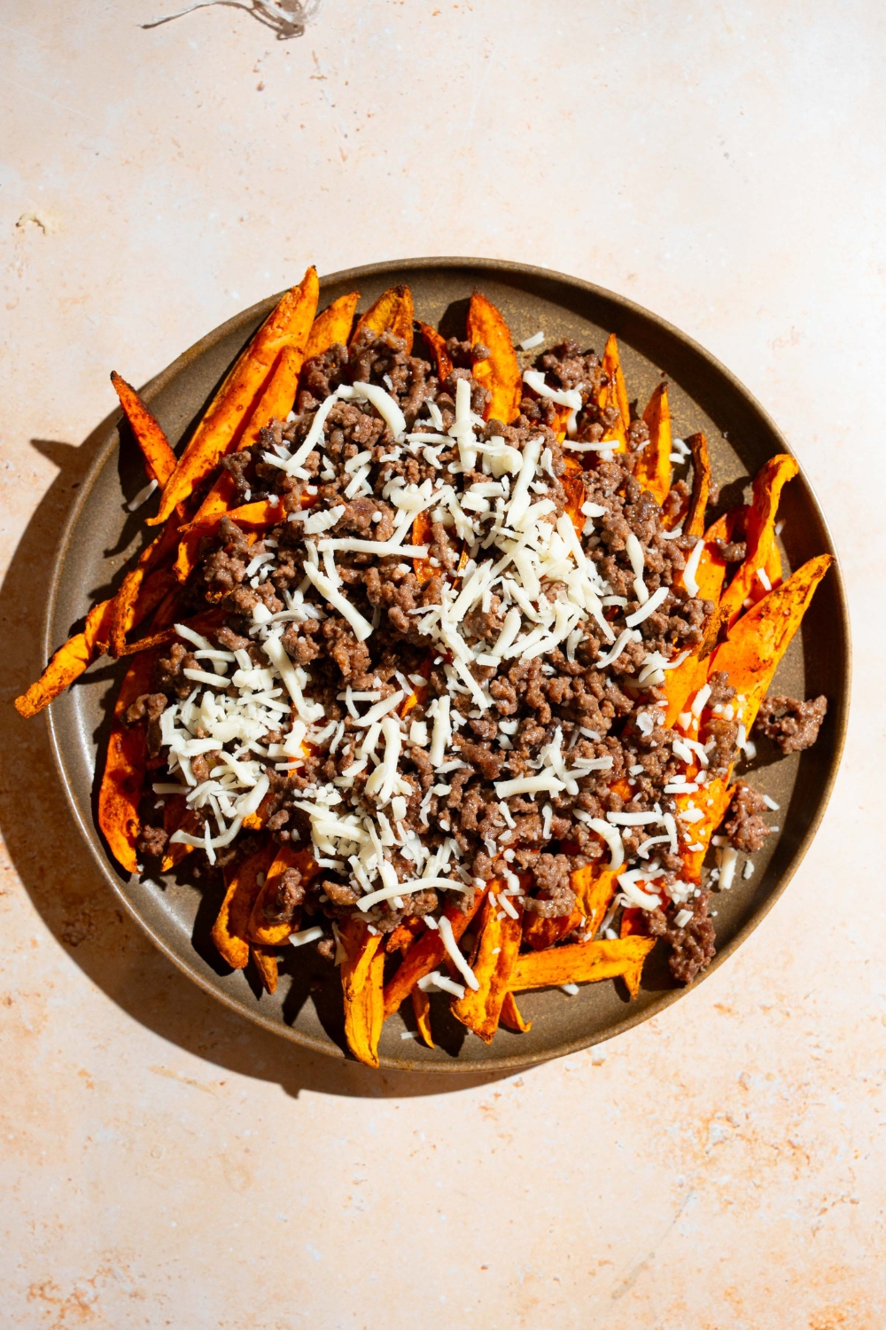 A plate with air fried sweet potato fries topped with ground beef and shredded cheese. The plate is on a tan counter.