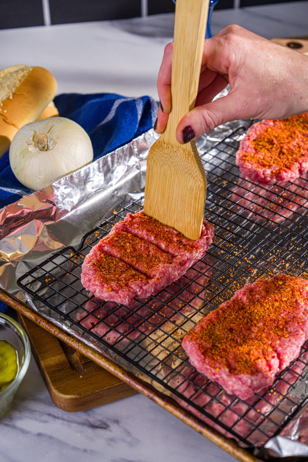 Several unbaked McRib patties topped with seasonings being scored with a wooden spoon. The patties are on a wire rack over a baking sheet lined with foil.