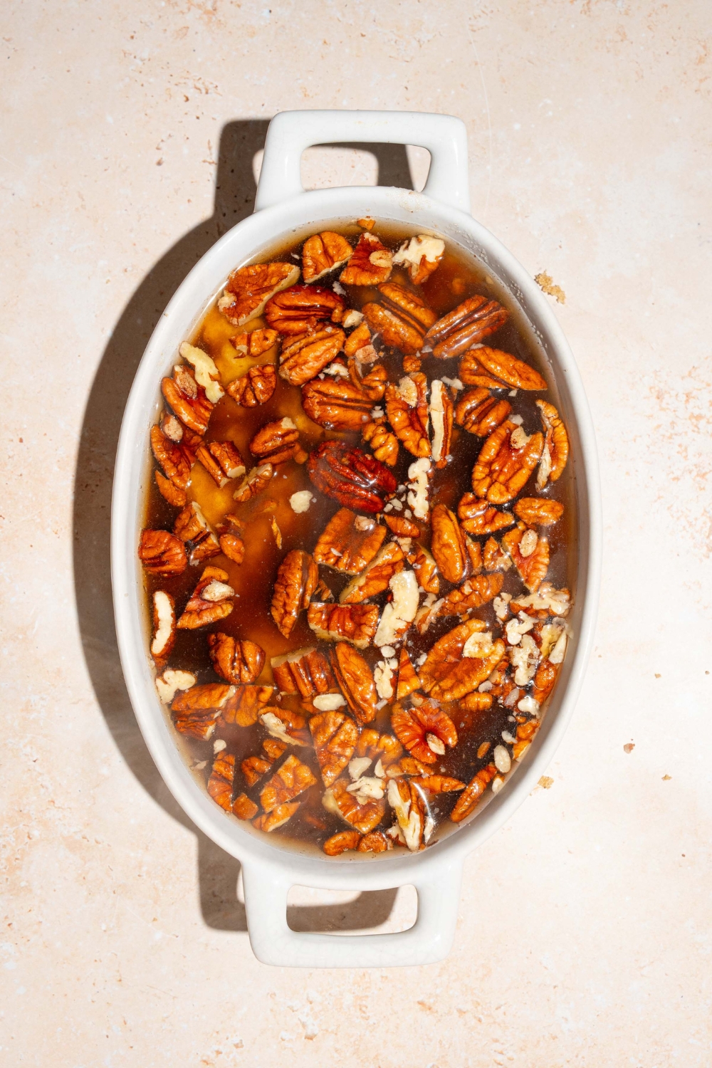 A casserole dish with pecan pie cobbler batter topped with pecans, brown sugar, and water. The dish is on a tan counter.