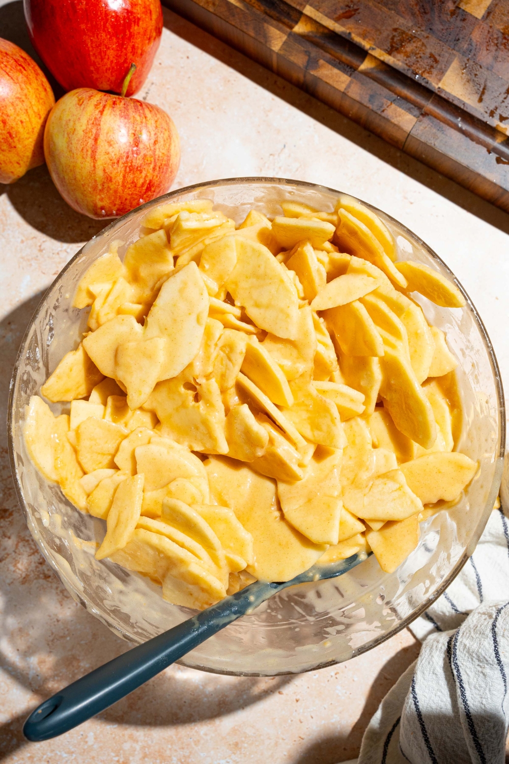 A large bowl with sliced apples tossed in a invisible apple cake batter. The bowl is on a tan counter with a wooden board and apples.