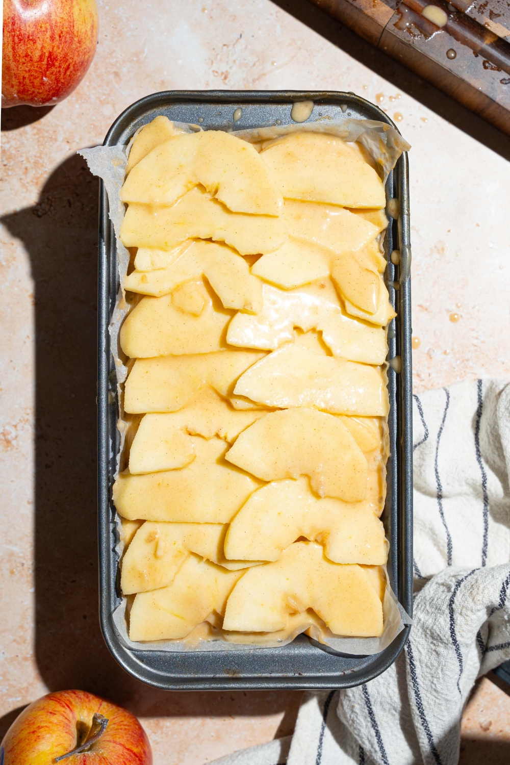 A loaf pan lined with parchment paper with unbaked invisible apple cake topped with sliced apples. The pan is on a tan counter with a white striped napkin and apples.