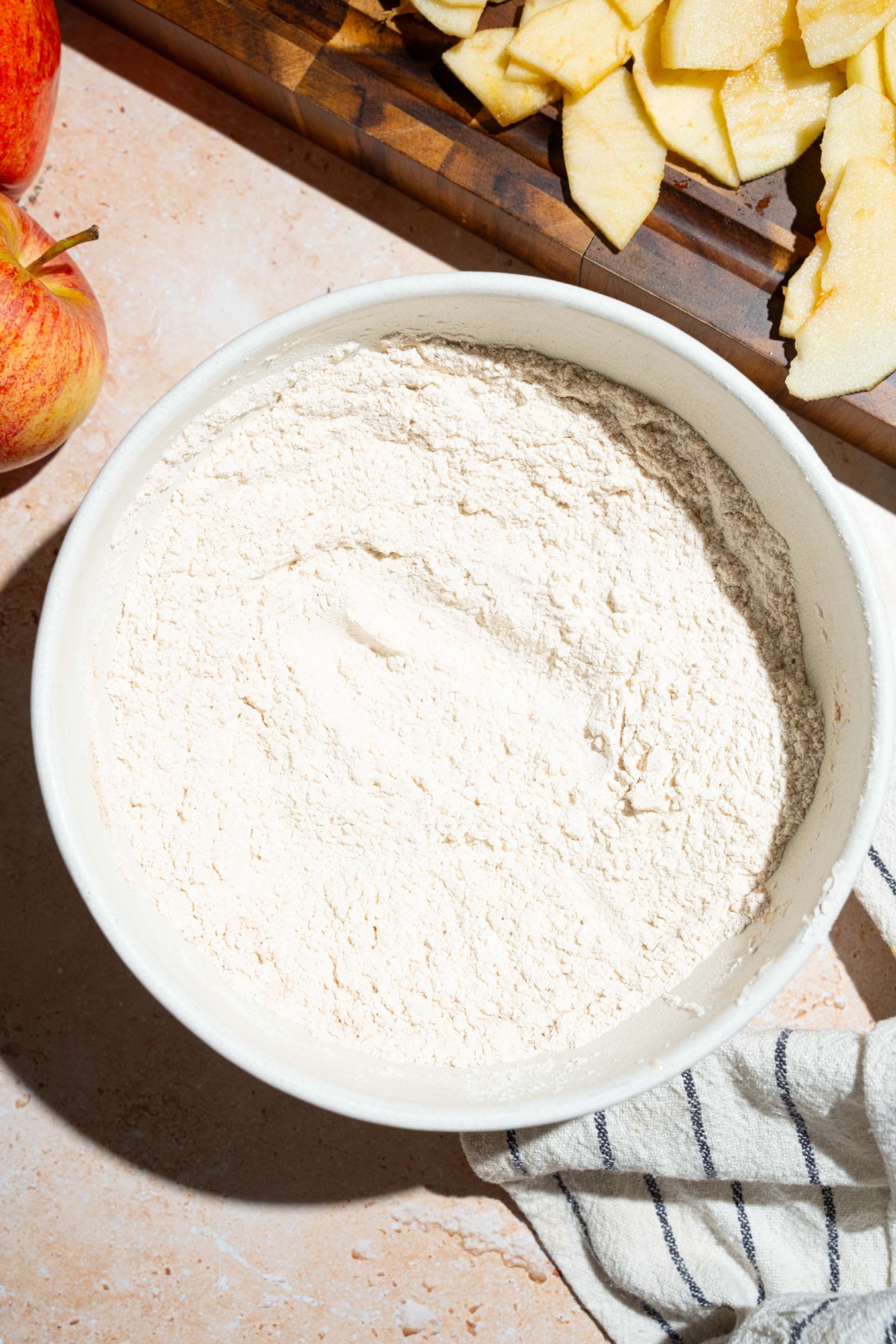 A large bowl with a mixture of dry ingredients to make invisible apple cake. The bowl is on a tan counter with a board of sliced apples.