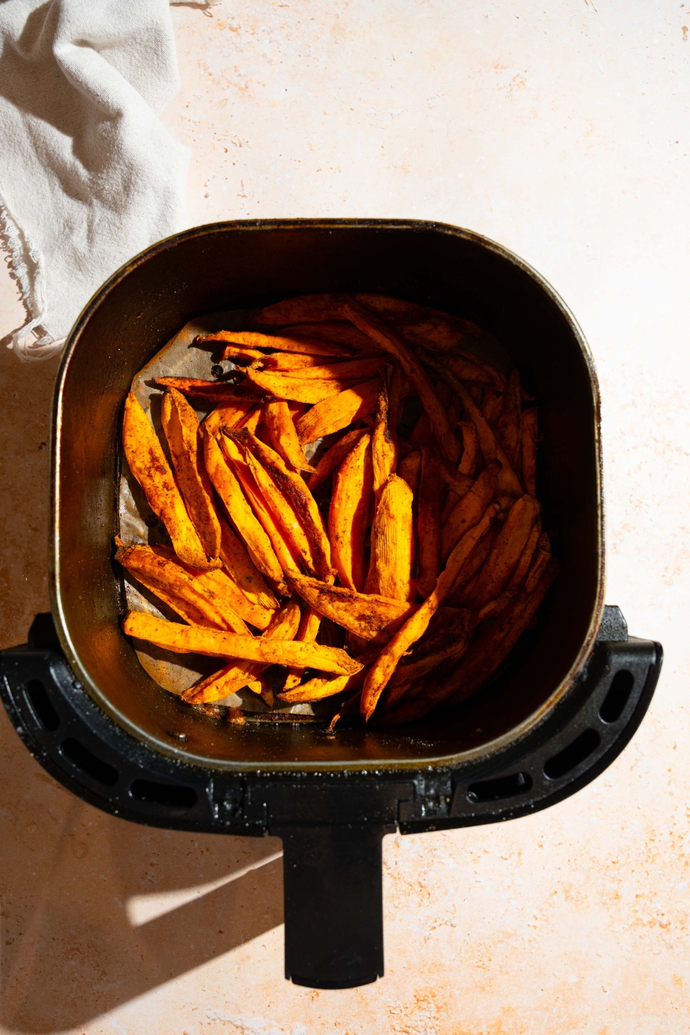 An air fryer basket with air fried seasoned sweet potato fries. The basket is on a tan counter.