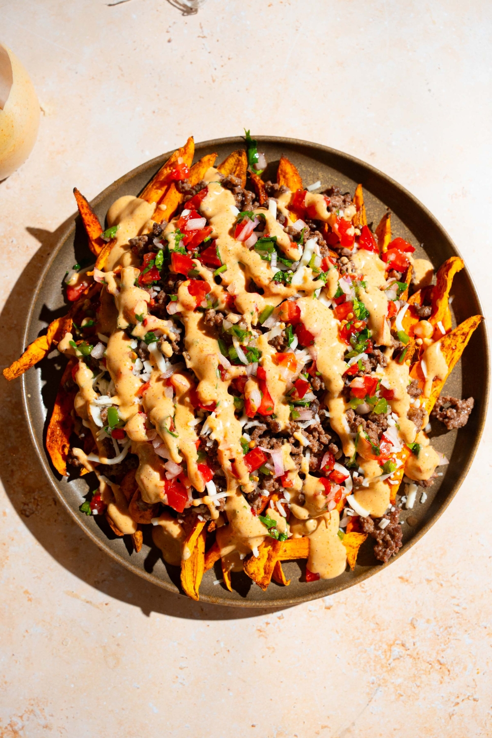 A plate of loaded sweet potato fries topped with ground beef, chipotle ranch, pico de gallo, and shredded cheese. The plate is on a tan counter.