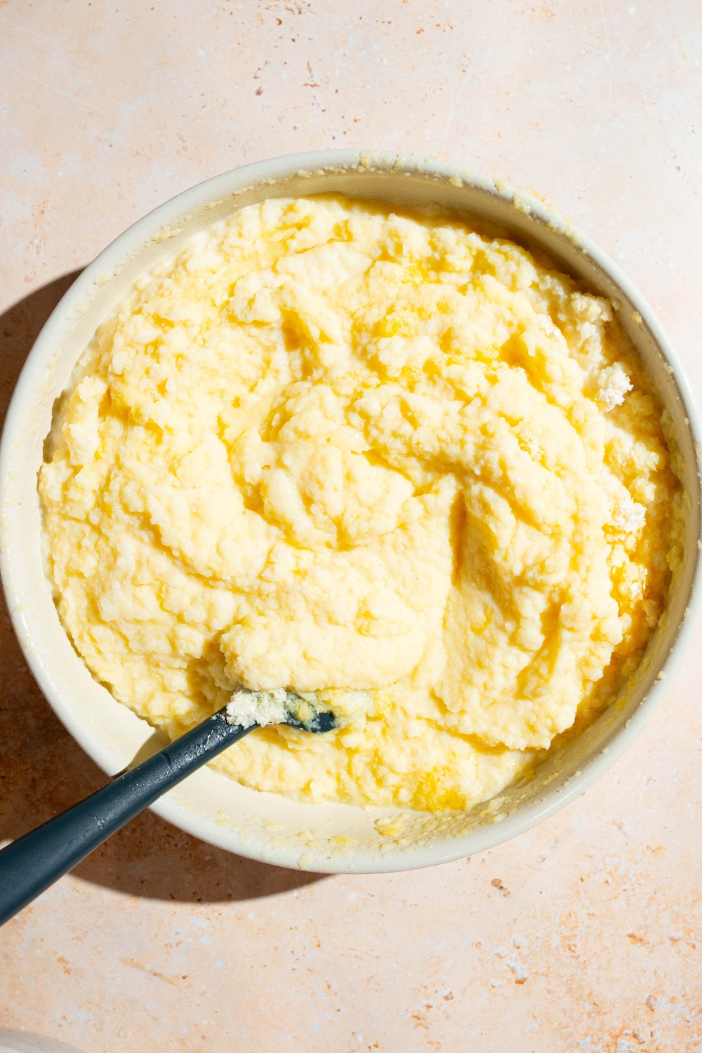 A large bowl with a rubber spoon mixing a mashed potato mixture with eggs and milk. The bowl is on a tan counter.