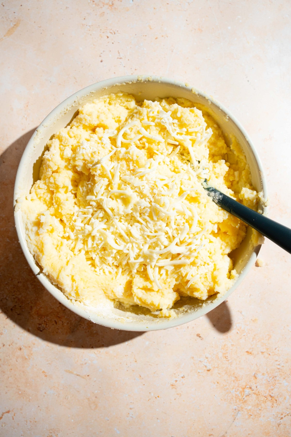 A large bowl with a mashed potatoes mixture topped with shredded cheese. A rubber spoon is in the bowl. The bowl is on a tan counter.