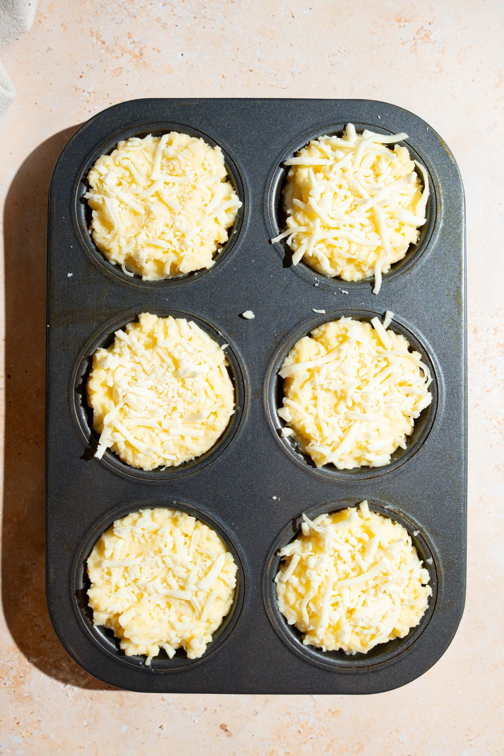 A muffin tin with mashed potato mixture in each cup topped with shredded cheese. The tin is on a tan counter.