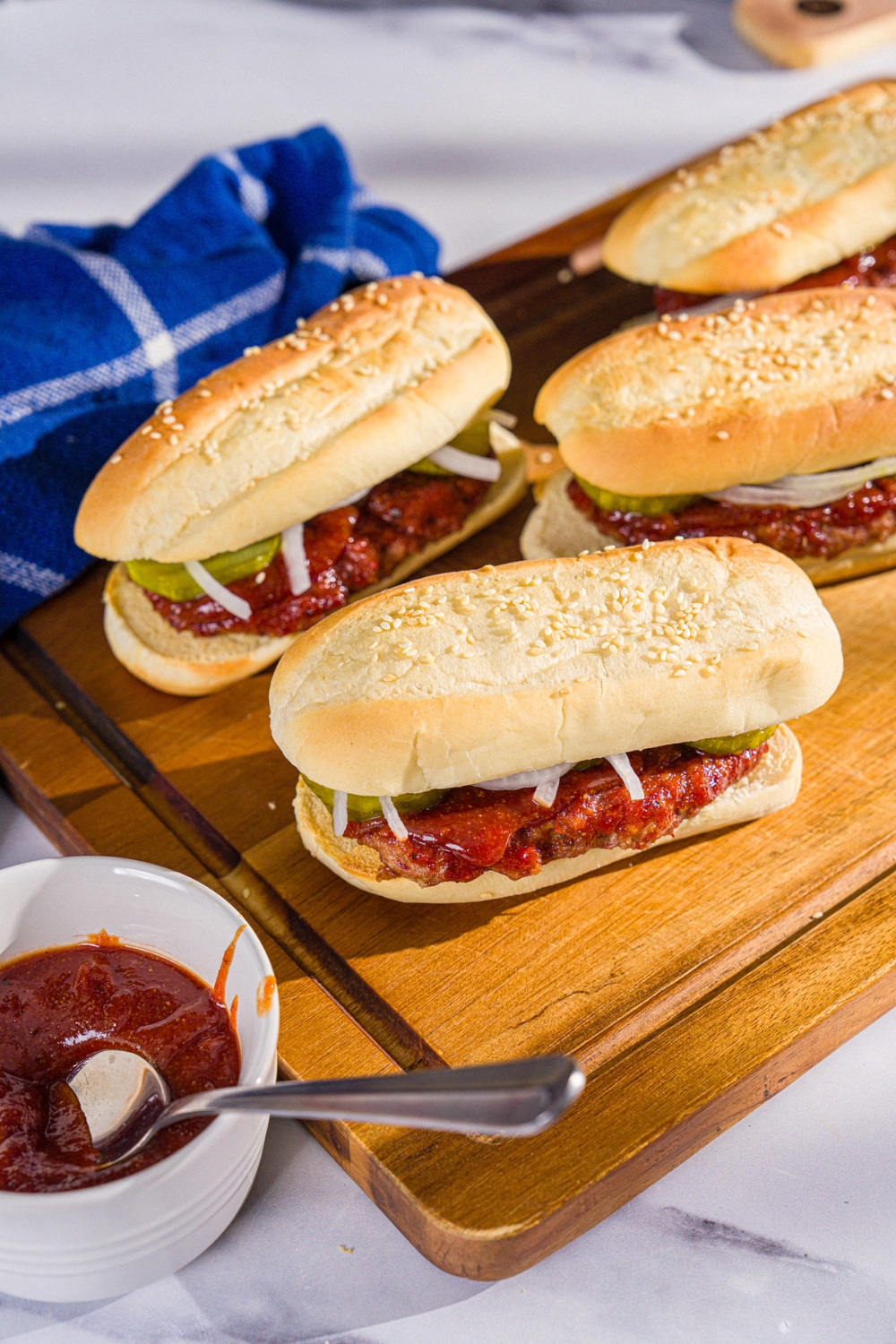 A wooden board with several homemade McRib sandwiches on a hoagie roll with pickles, onions, and homemade BBQ sauce. The board is on a marble counter with a bowl of BBQ sauce.