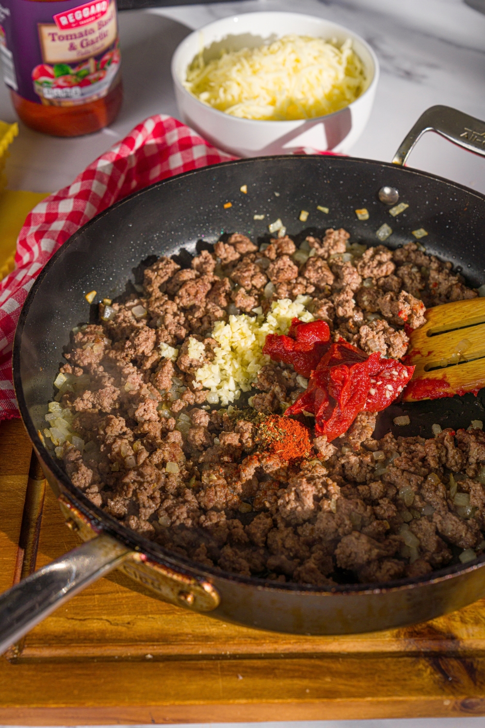A skillet with browned ground beef topped with tomato paste and garlic. A wooden spoon is stirring the mixture. The skillet is on a wooden board.