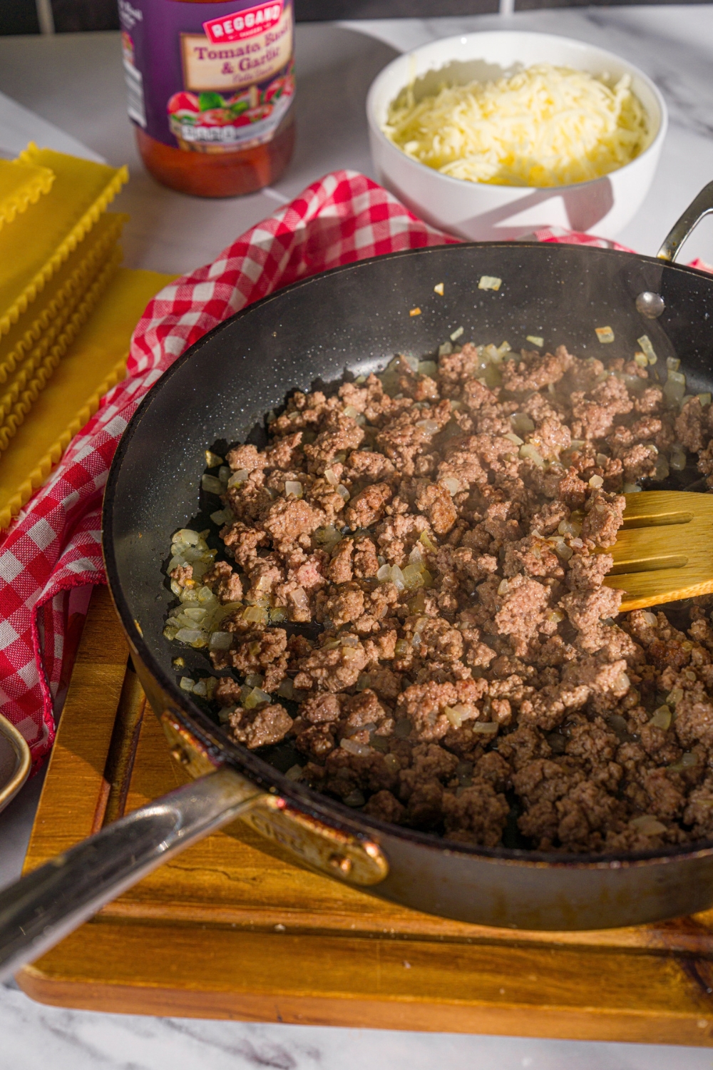 A skillet with browned ground beef and onion. There is a wooden spoon stirring the mixture. The skillet is on a wooden board.