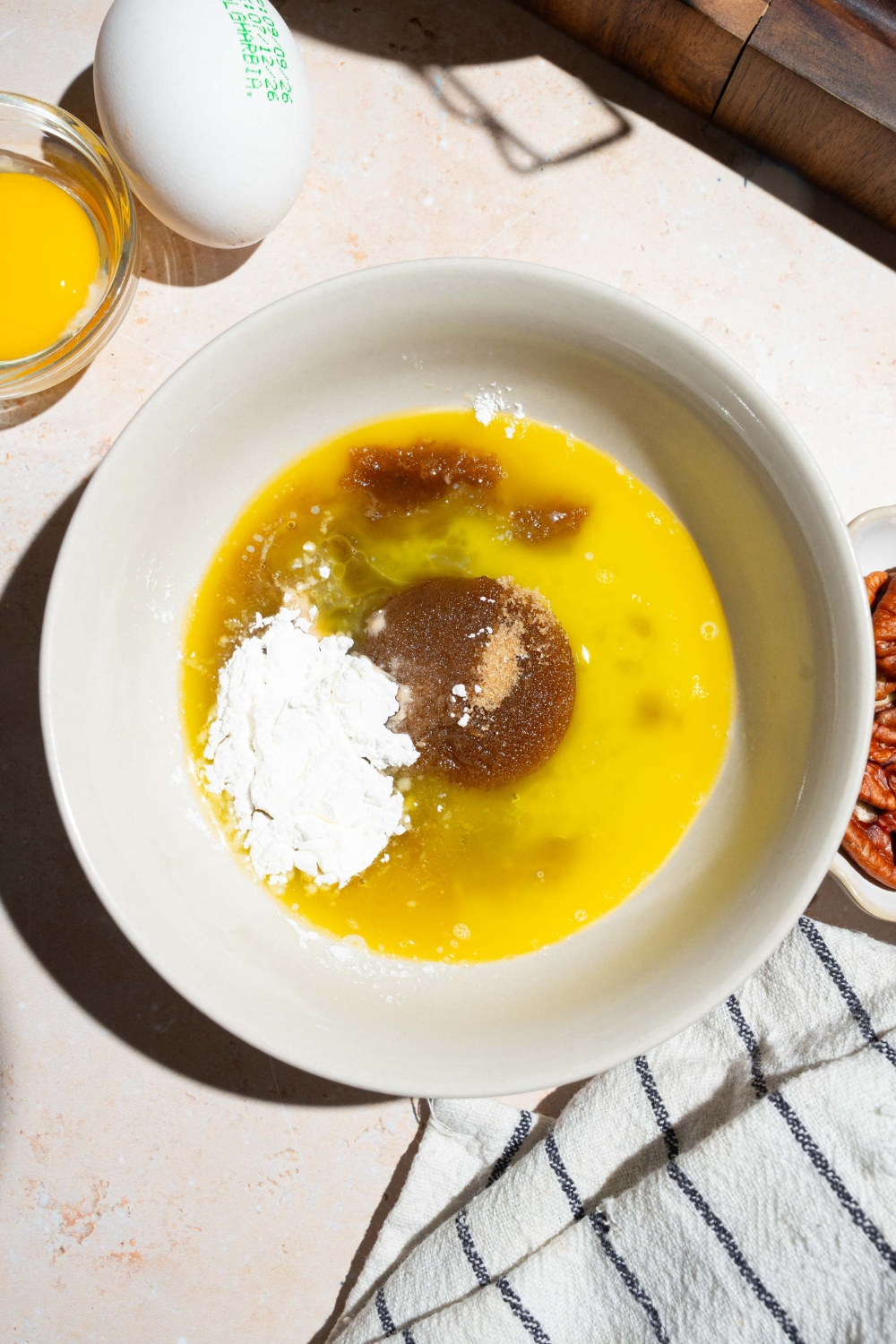 A bowl with melted butter, sugar, egg, vanilla, and brown sugar. The bowl is on a tan counter with a white striped napkin.