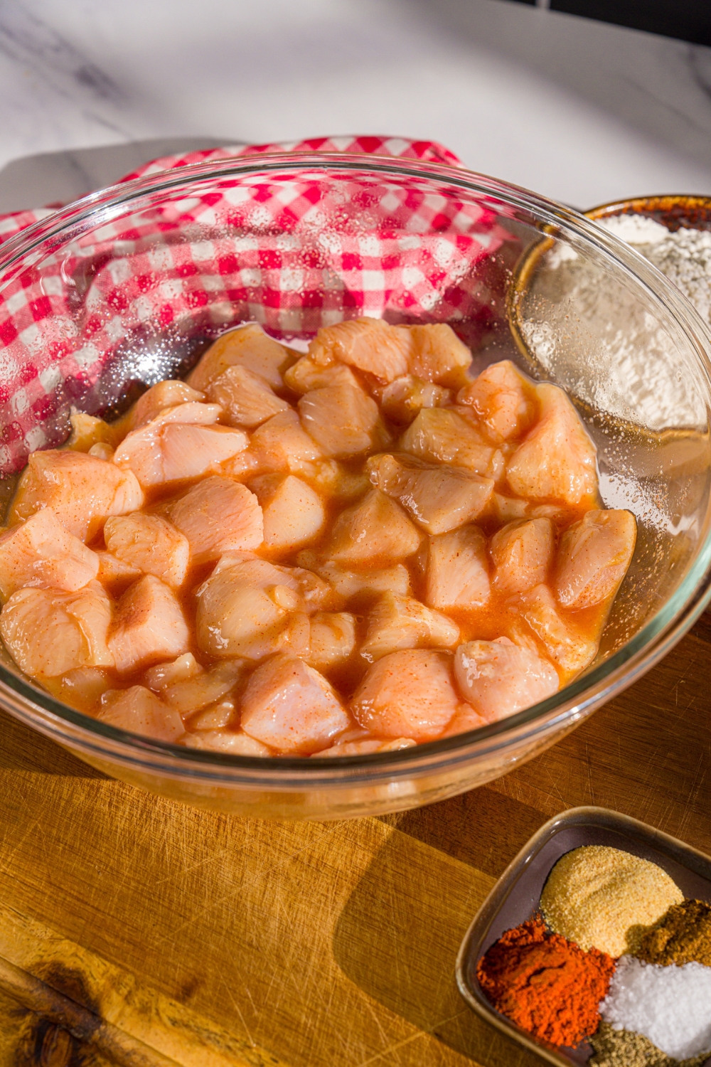 A large bowl of chicken pieces marinating in seasonings and club soda. The bowl is on a wooden board with a small bowl of seasonings.