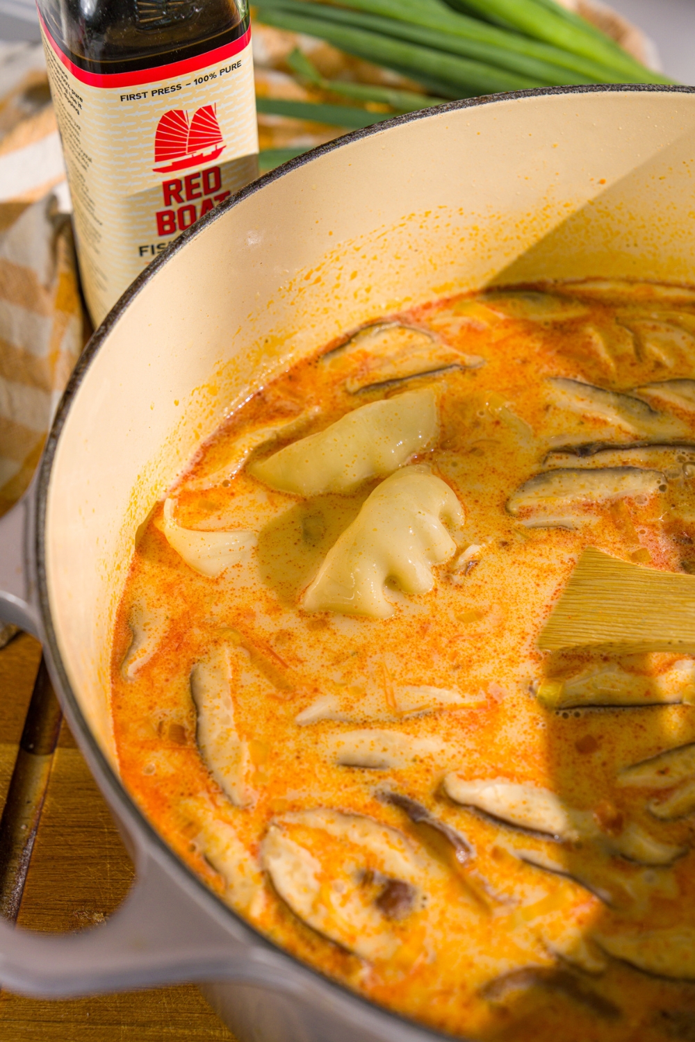A dutch oven with coconut red curry broth with dumplings. There is a wooden spoon stirring the soup.
