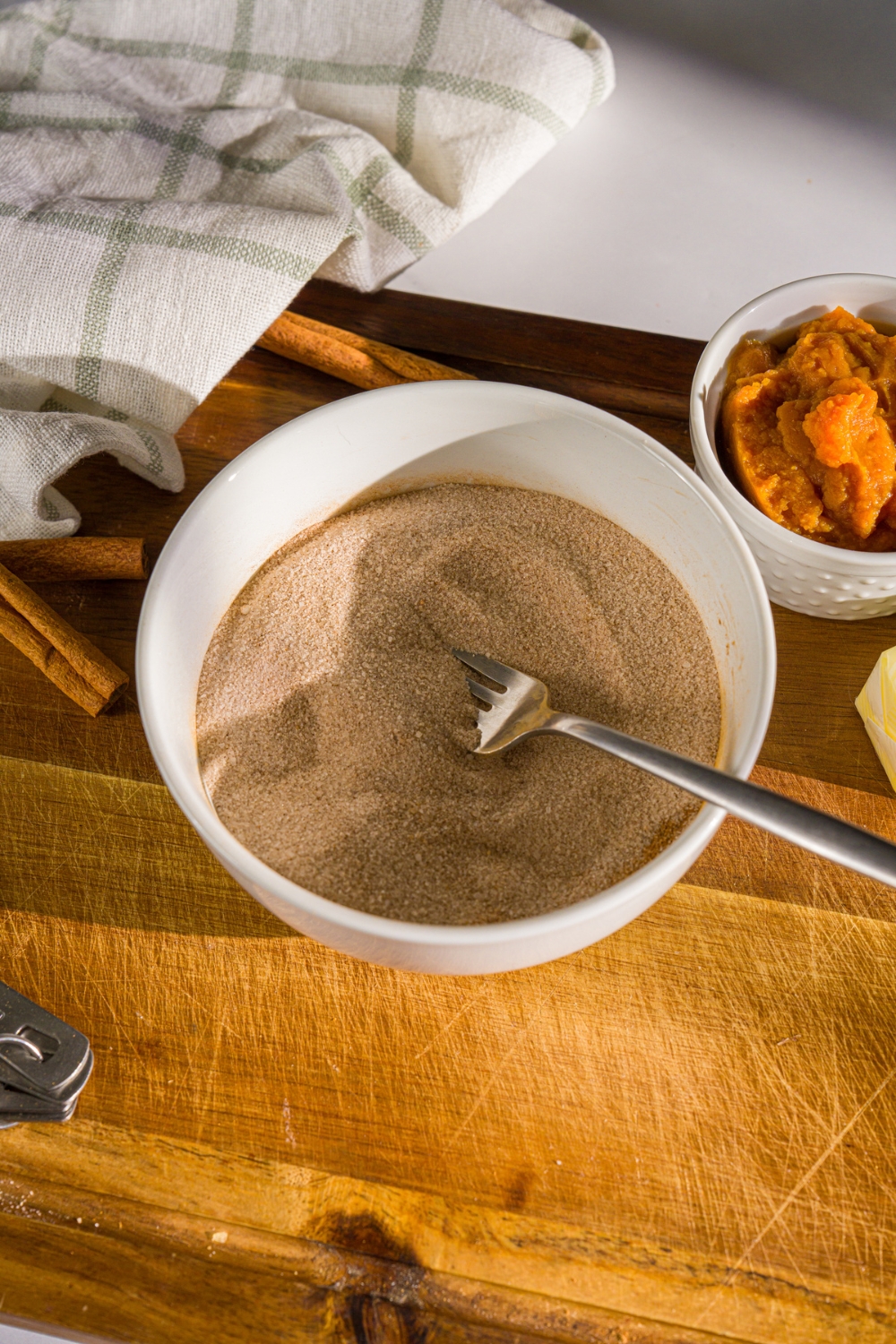 A white bowl with a cinnamon sugar mixture with a fork in the bowl. The bowl is on a wooden board with a bowl of pumpkin puree and white cloth napkin.