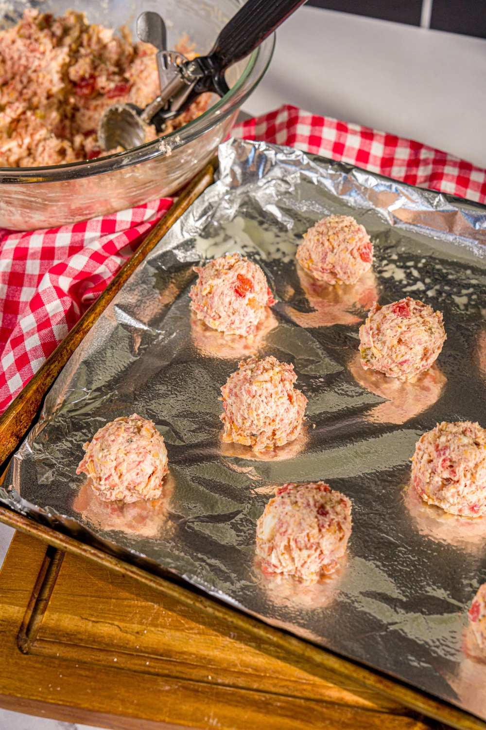 A baking sheet lined with foil with shaped uncooked cheddar bay biscuit sausage rolls. The sheet is on a wooden board with a red checkered napkin.