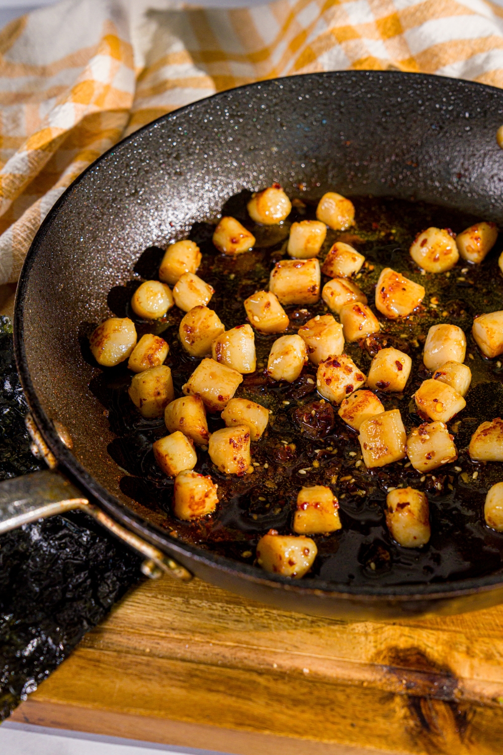 A skillet with scallops seared in oil. The skillet is on a wooden board with a yellow cloth napkin.