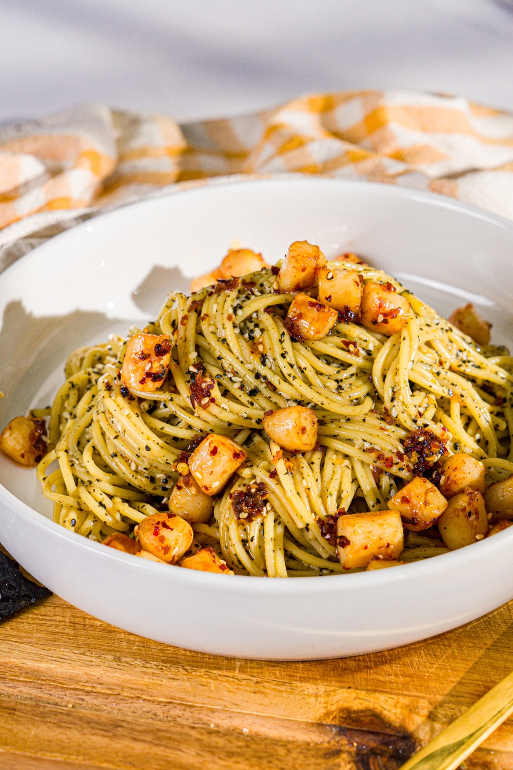 A bowl of seaweed pasta topped with seared scallops and chili crisp. The bowl is on a wooden board.