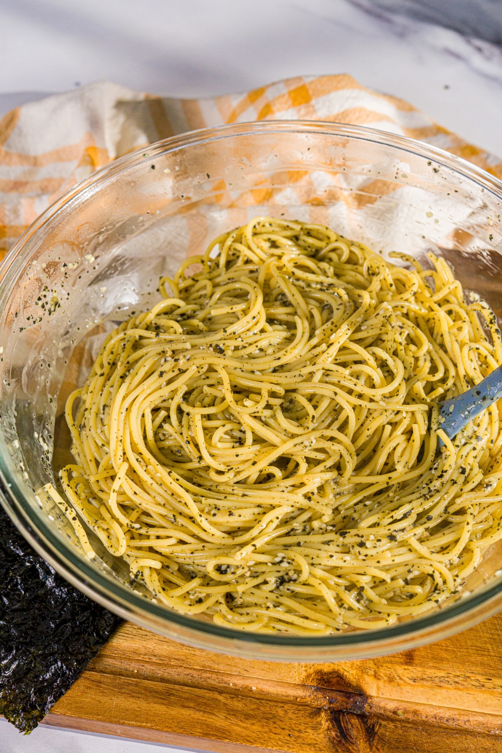 A large bowl with a rubber spoon stirring a spaghetti and seaweed sauce mixture. The bowl is on a wooden board with a yellow cloth napkin.