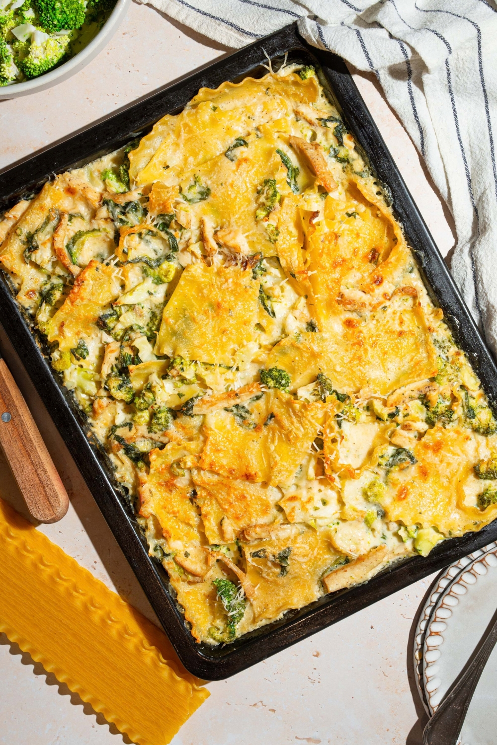 A sheet pan with chicken broccoli lasagna. The pan is on a tan counter with a white cloth napkin and bowl of broccoli.