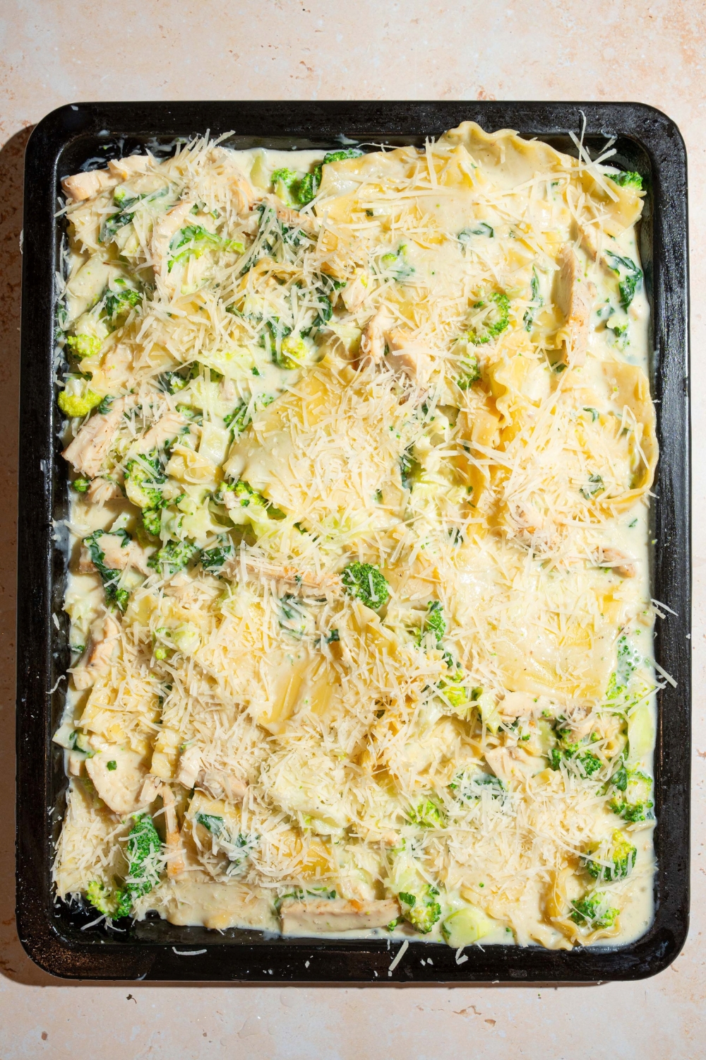 A sheet pan with a chicken broccoli lasagna mixture topped with shredded cheese. The pan is on a tan counter.