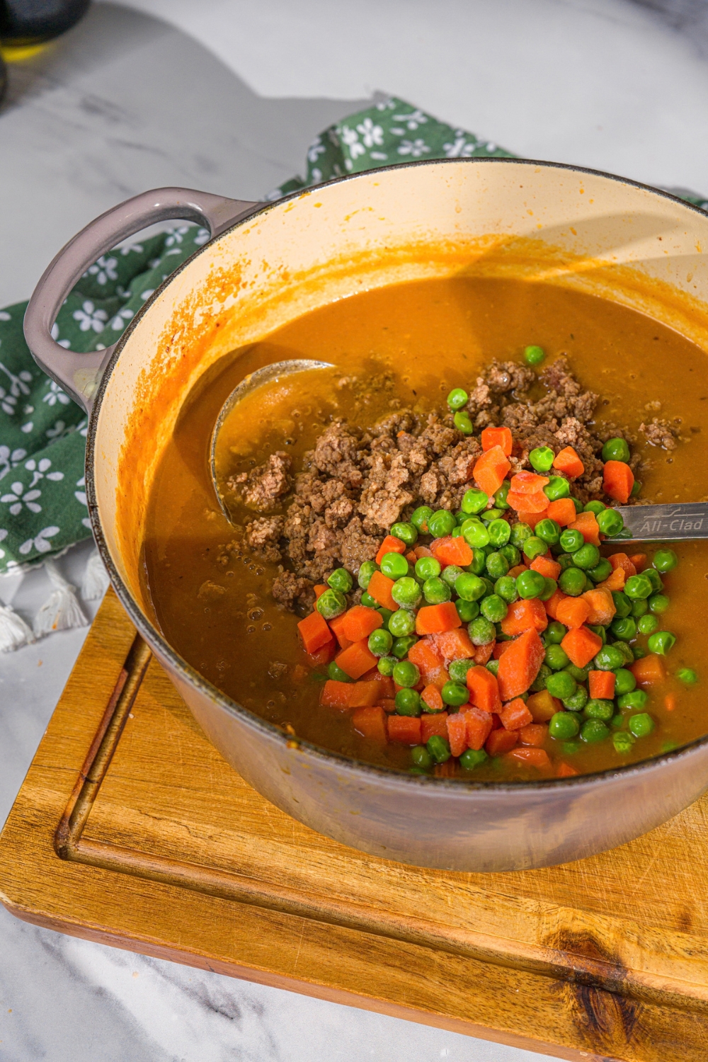 A large pot with ground beef, carrots, and peas being added to a shepherd's pie soup base. There is a ladle in the pot. The pot is on a wooden board.