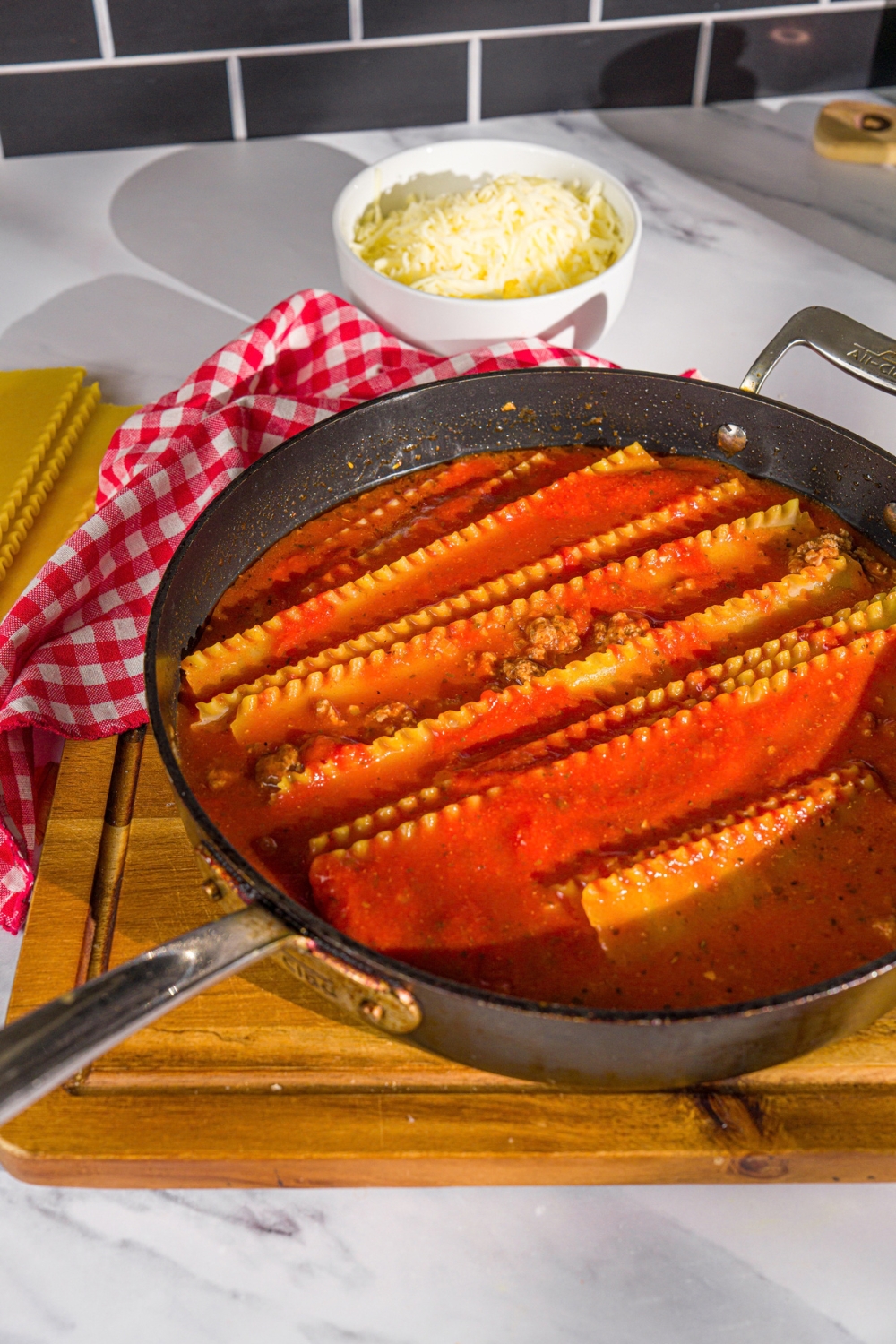 A skillet with uncooked lasagna noodles in tomato sauce. The skillet is on a wooden board with a red checkered napkin and small bowl of shredded cheese.