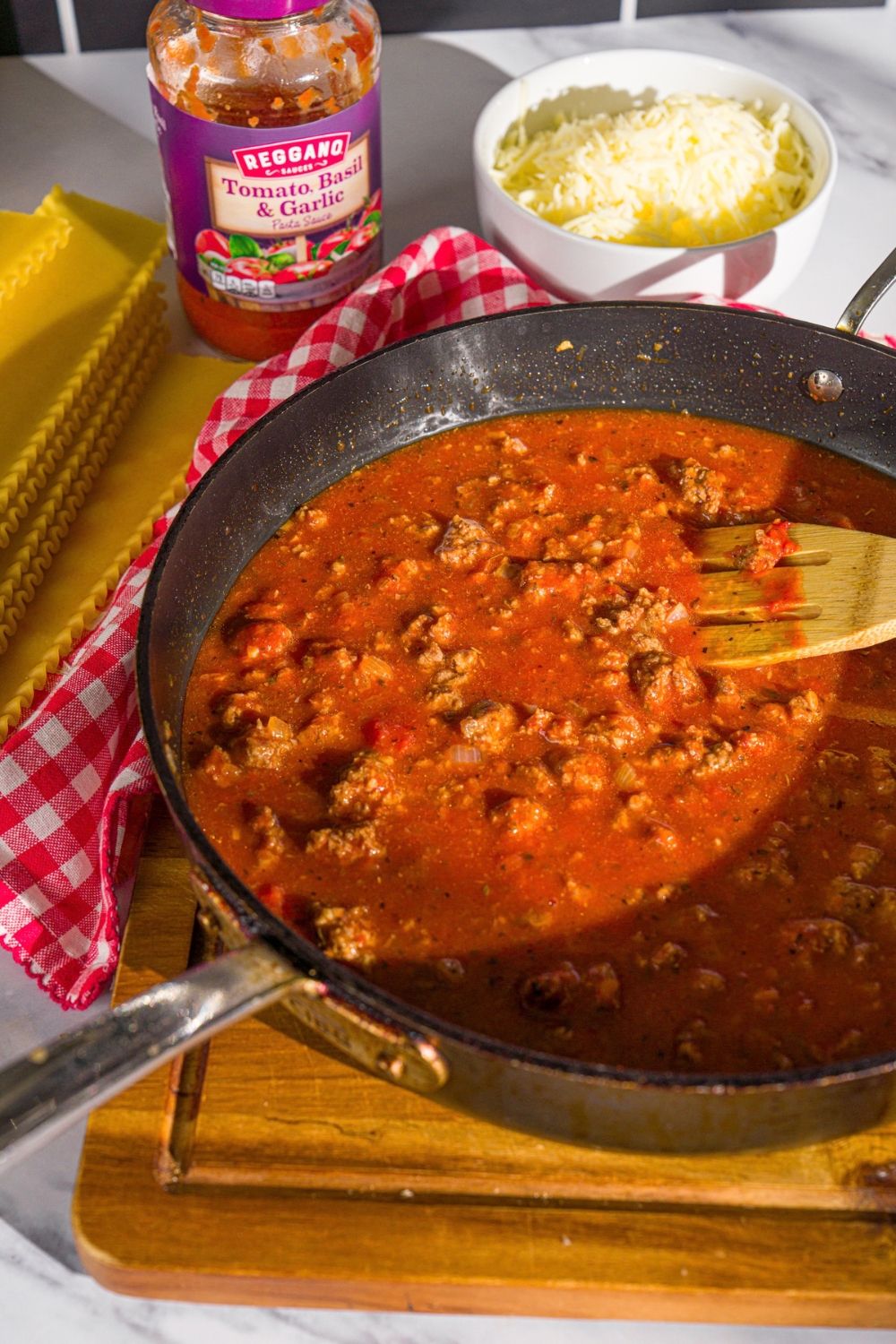 A skillet with ground beef cooking in tomato sauce. The skillet is on a wooden board. A wooden spoon is stirring the mixture.