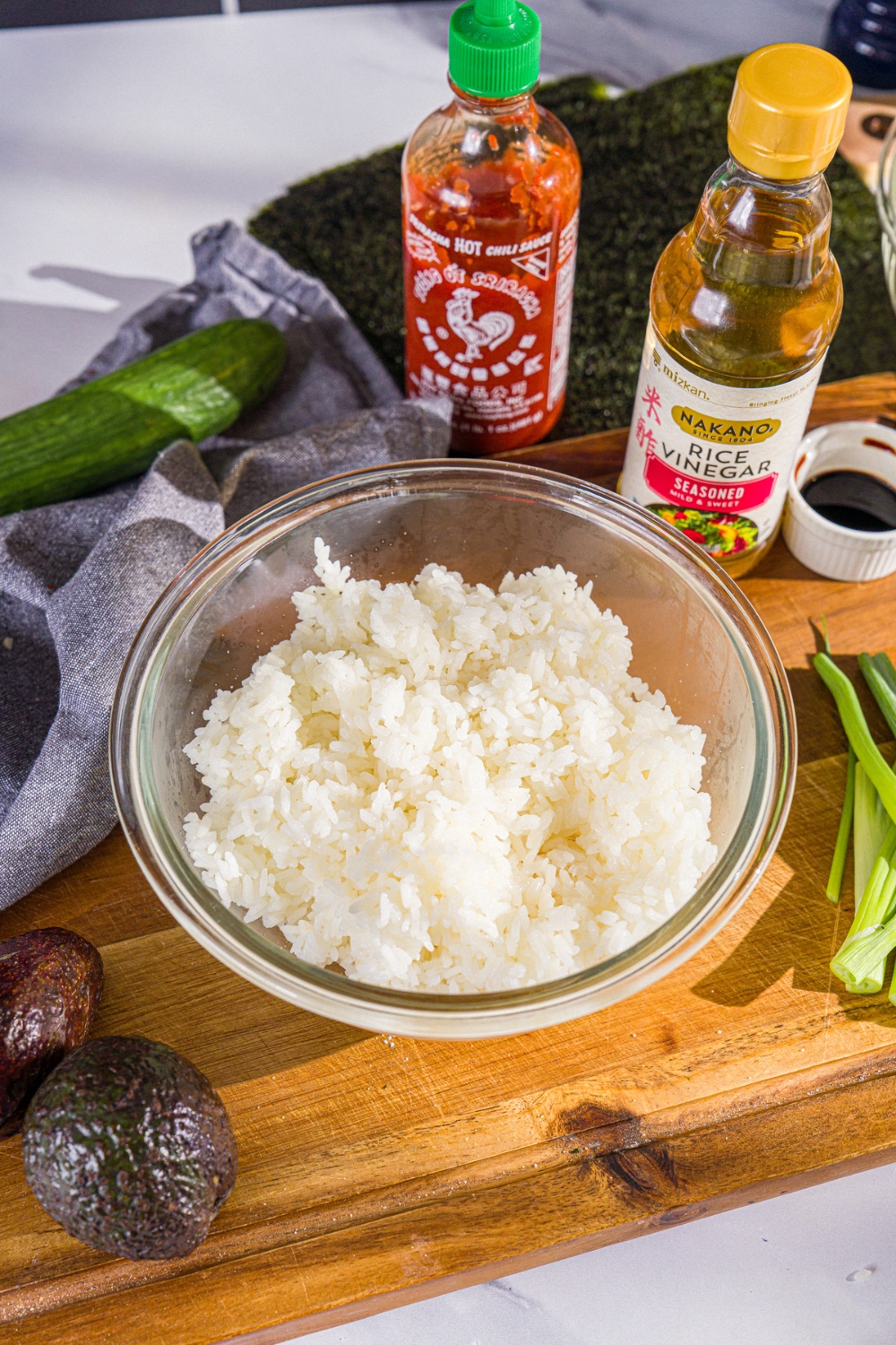 A glass bowl of sushi rice. The bowl is on a wooden board with rice vinegar, sriracha, avocado, and green onions.