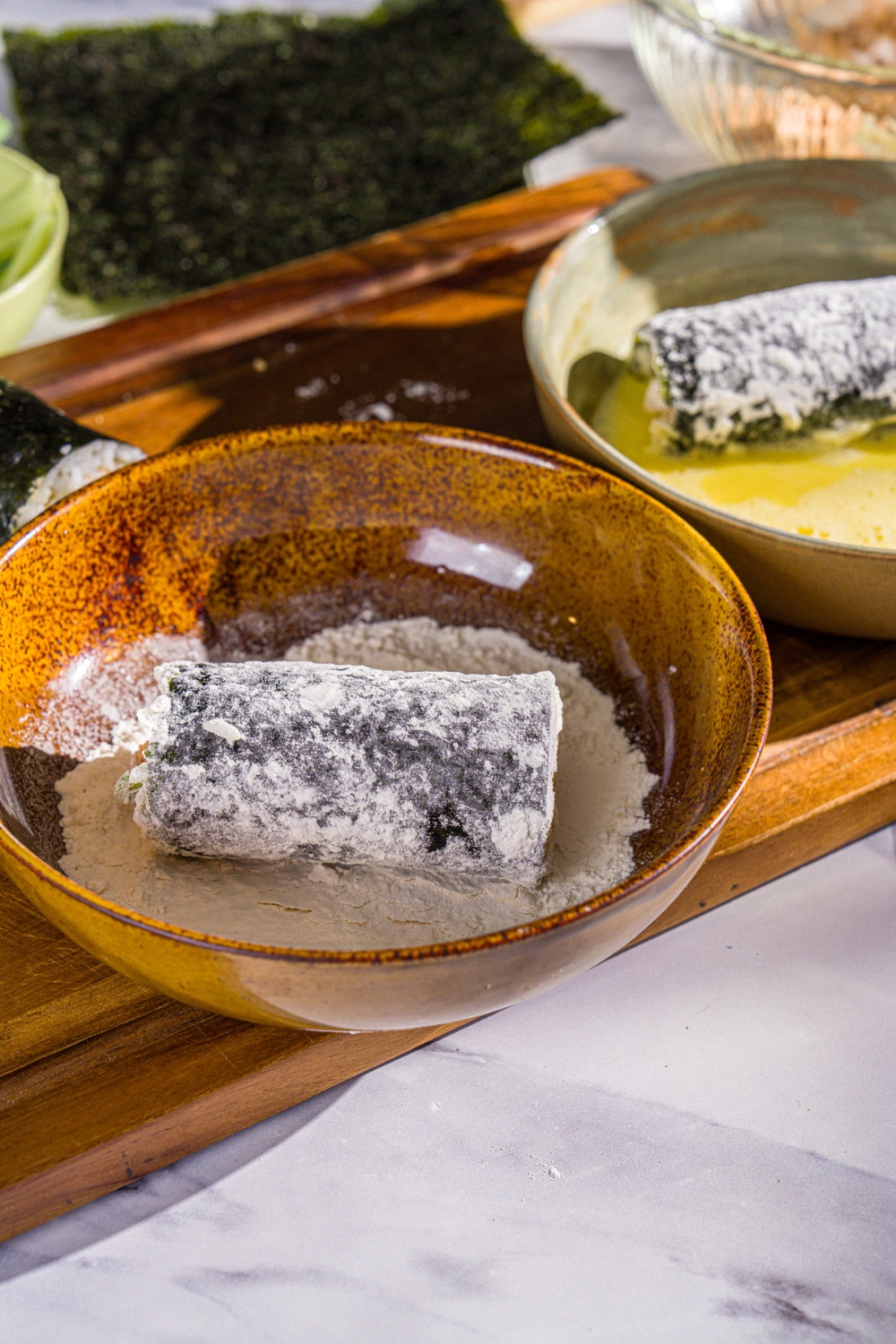 A sliced sushi roll in a bowl of flour. The bowl is on a wooden board with a bowl of whisked egg.