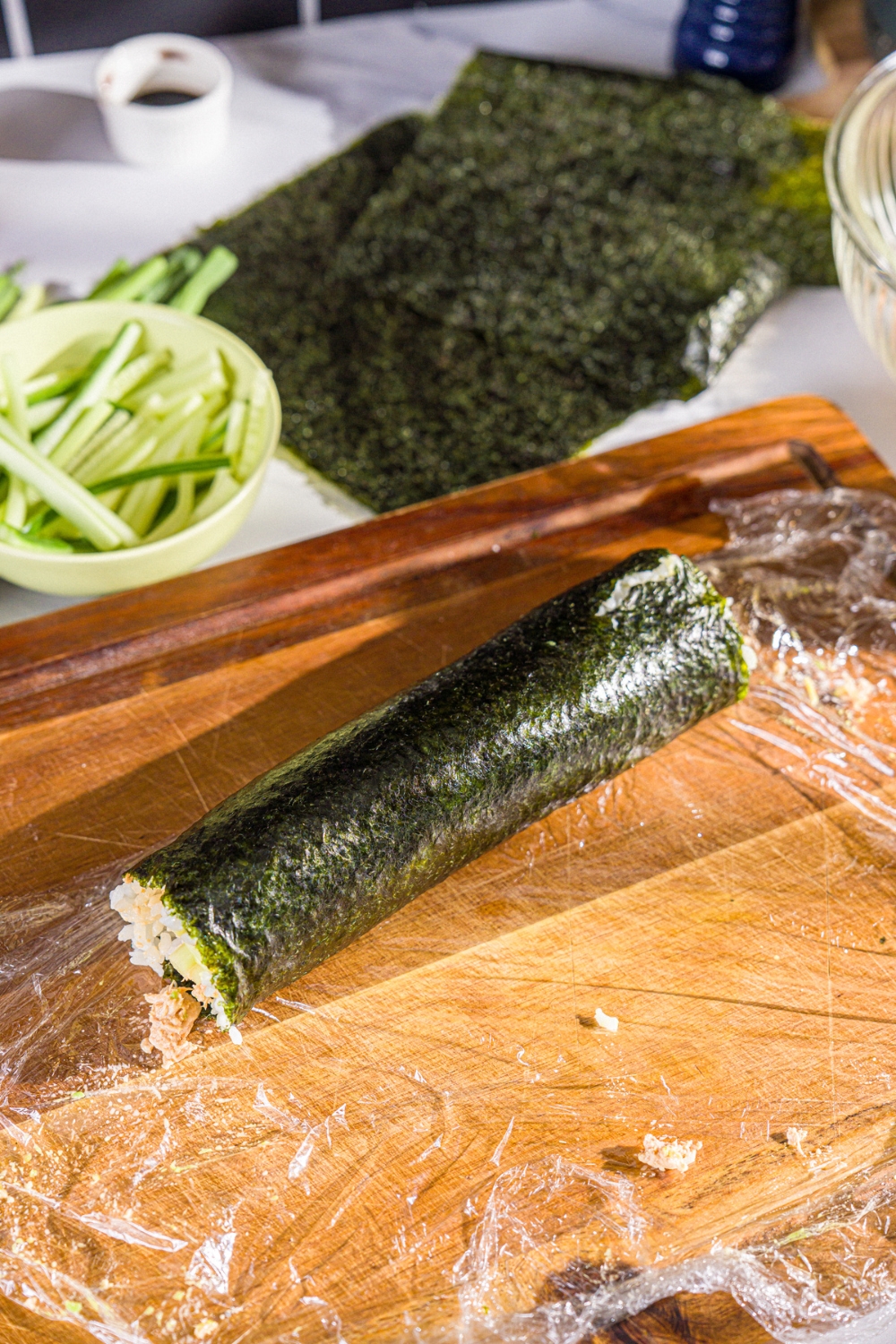 A wooden board with a rolled tuna sushi burrito rolled in saran wrap. The board is on a marble counter with a bowl of cucumber and seaweed sheets.