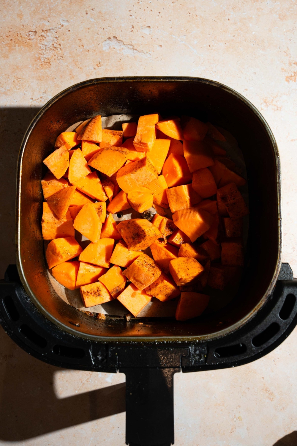 An air fryer basket with cubed seasoned sweet potatoes. The basket is on a tan counter.