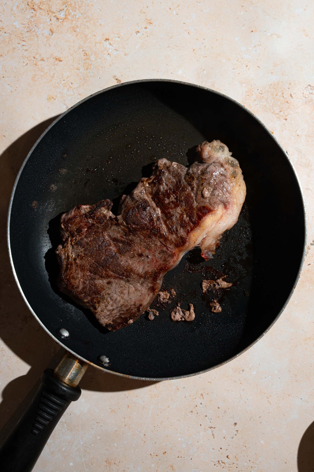 A skillet with seared seasoned steak. The skillet is on a tan counter.