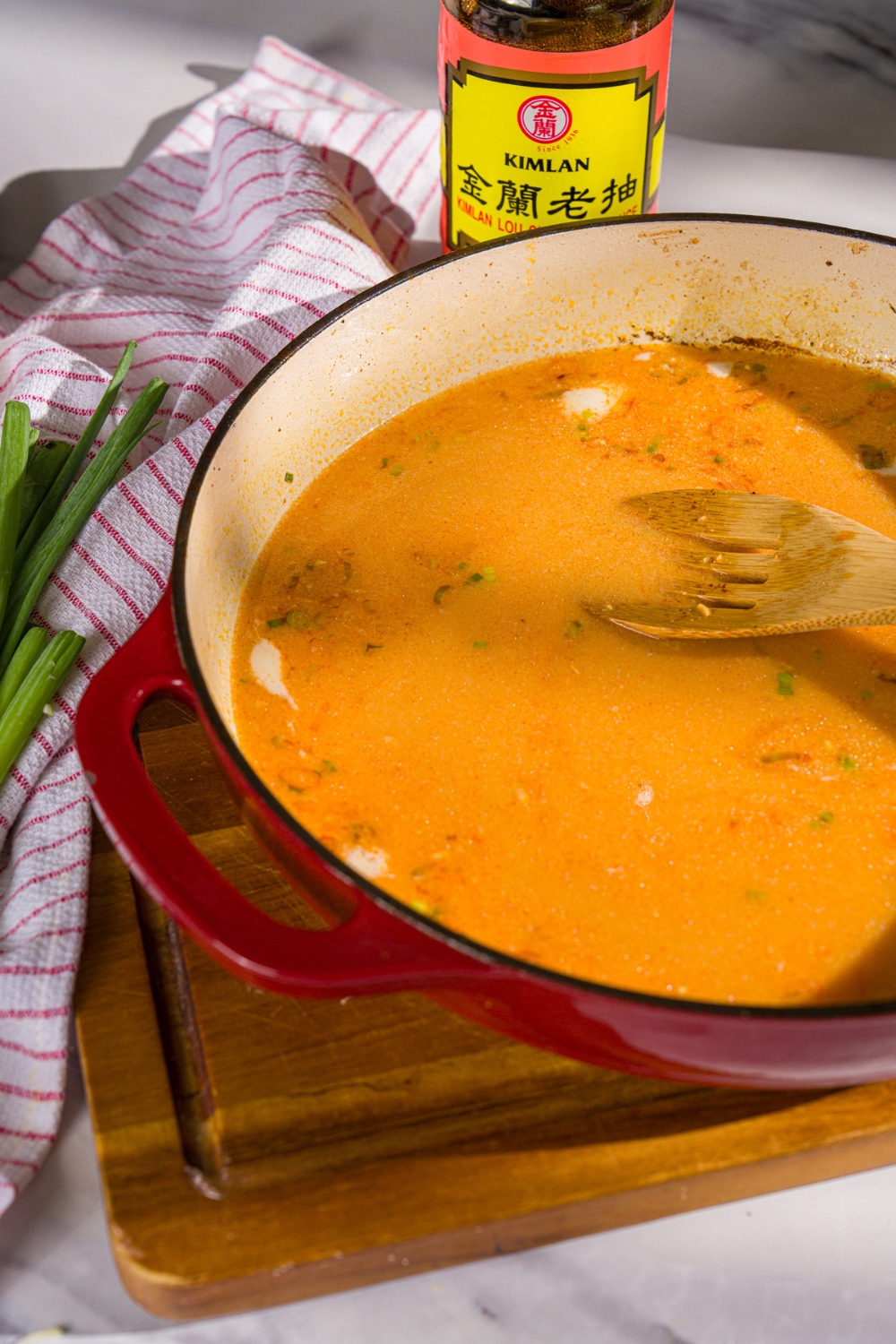 A skillet with a thai coconut chicken broth. A wooden spoon is stirring the broth. The skillet is on a wooden board.