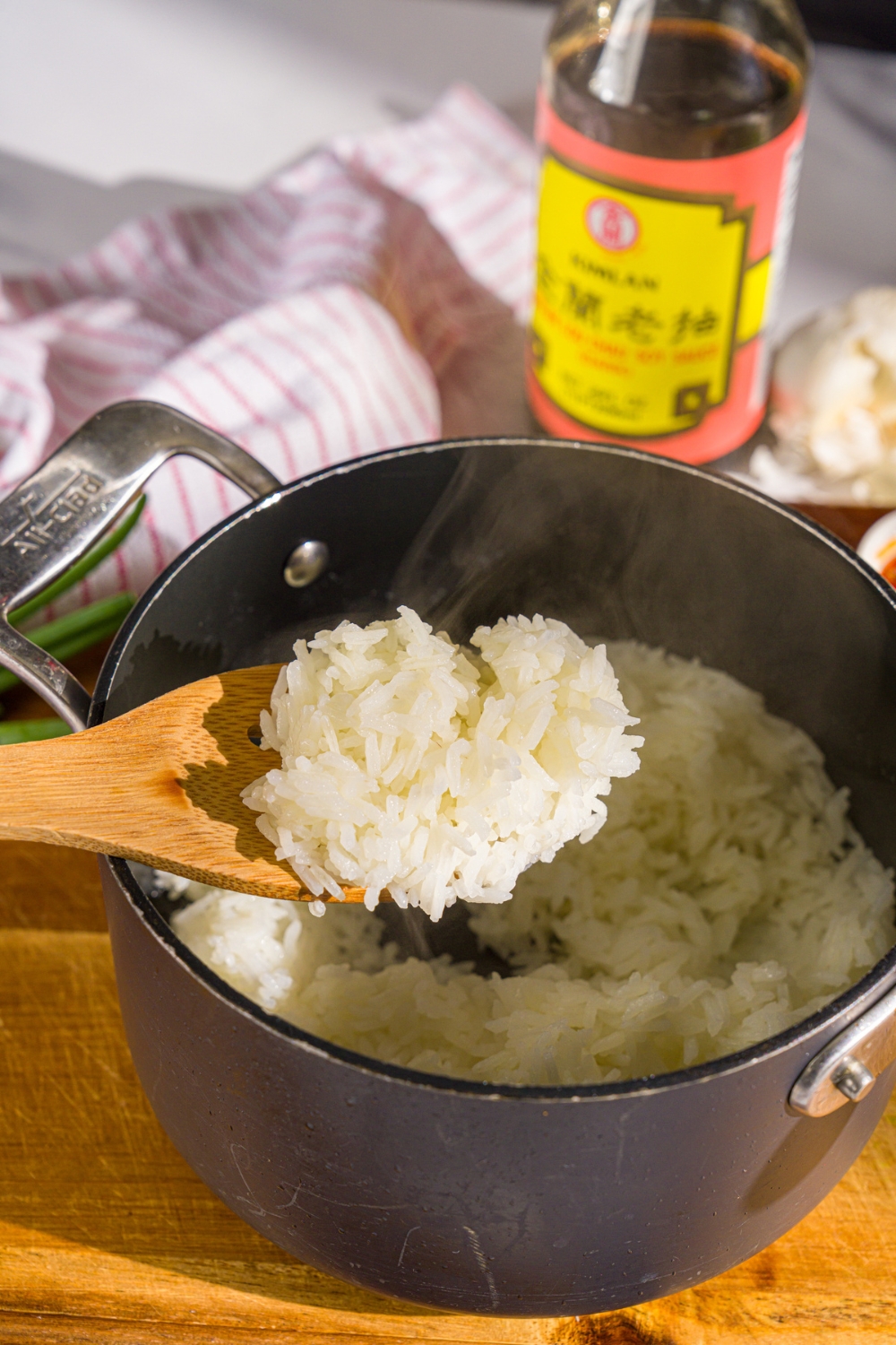A small pot with cooked jasmine rice. A wooden spoon is scooping some rice. The pot is on a wooden board.