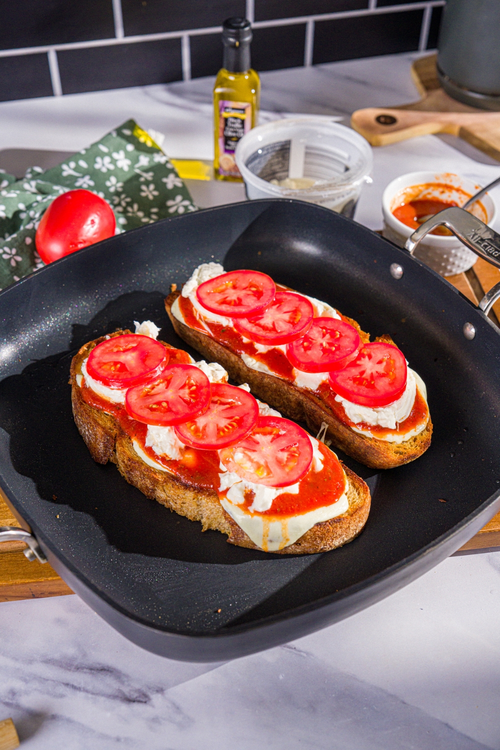 A skillet with two pieces of baked pizza toast with sourdough bread, mozzarella cheese, marinara sauce, burrata, and tomatoes. The skillet is on a wooden board.