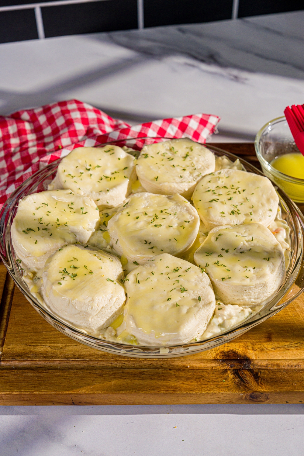 A round baking dish with chicken pot pie soup mixture topped with unbaked split biscuits covered with melted butter. The dish is on a wooden board with a red checkered napkin.