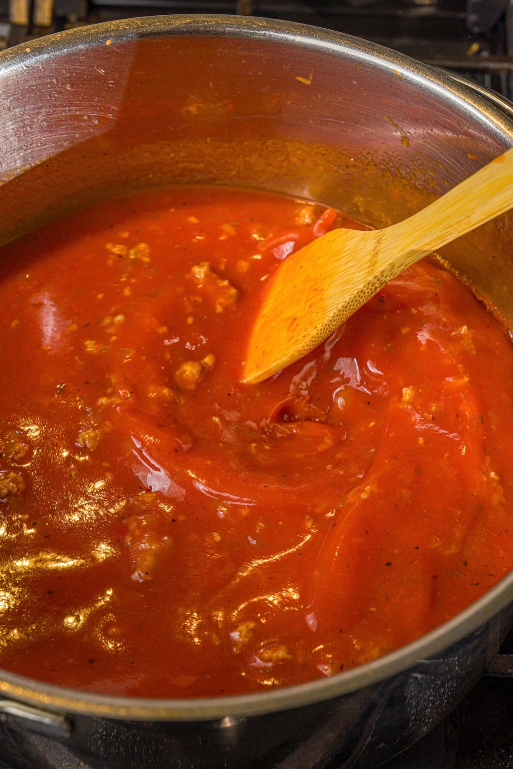 A large pot with marinara sauce and chicken stock with ground sausage and onion. A wooden spoon is stirring the mixture. The pot is cooking on the stove.