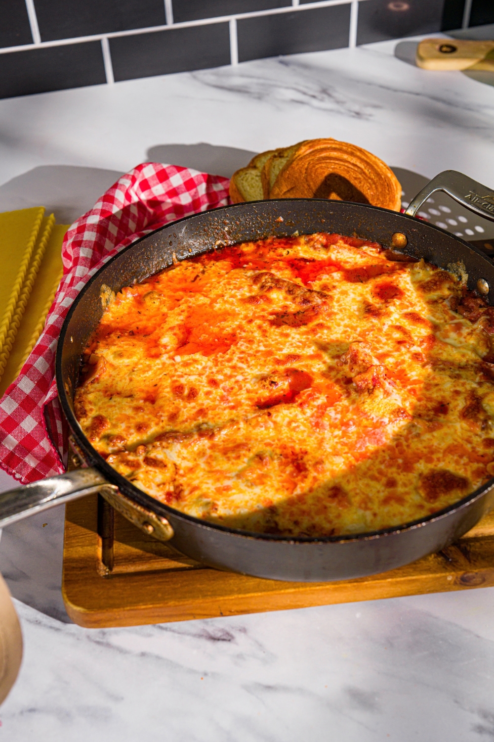 A skillet with cooked lasagna with ground beef, tomato sauce, and melted cheese on top. The skillet is on a wooden board with a red checkered napkin.