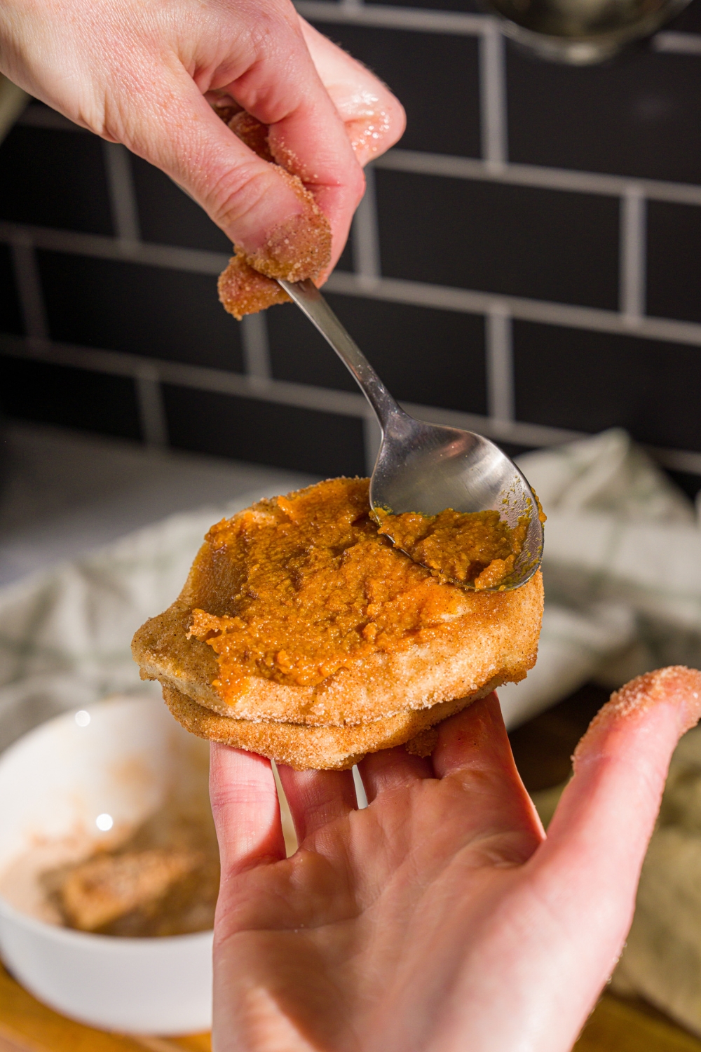 A spoon spreading pumpkin puree on a split biscuit coated in cinnamon sugar. There is a bowl of cinnamon sugar blurred in the background.
