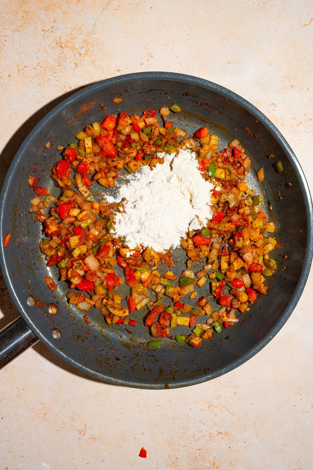 A skillet with sautéed vegetables tossed in cajun mixture topped with flour. The skillet is on a tan counter.