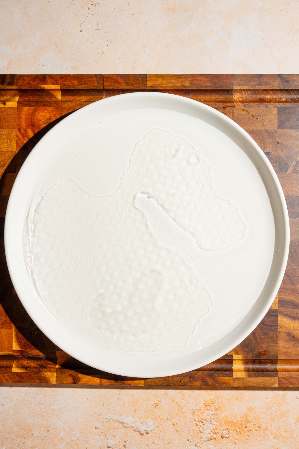 A white plate with rice paper sheet soaking in water. The plate is on a wooden board.