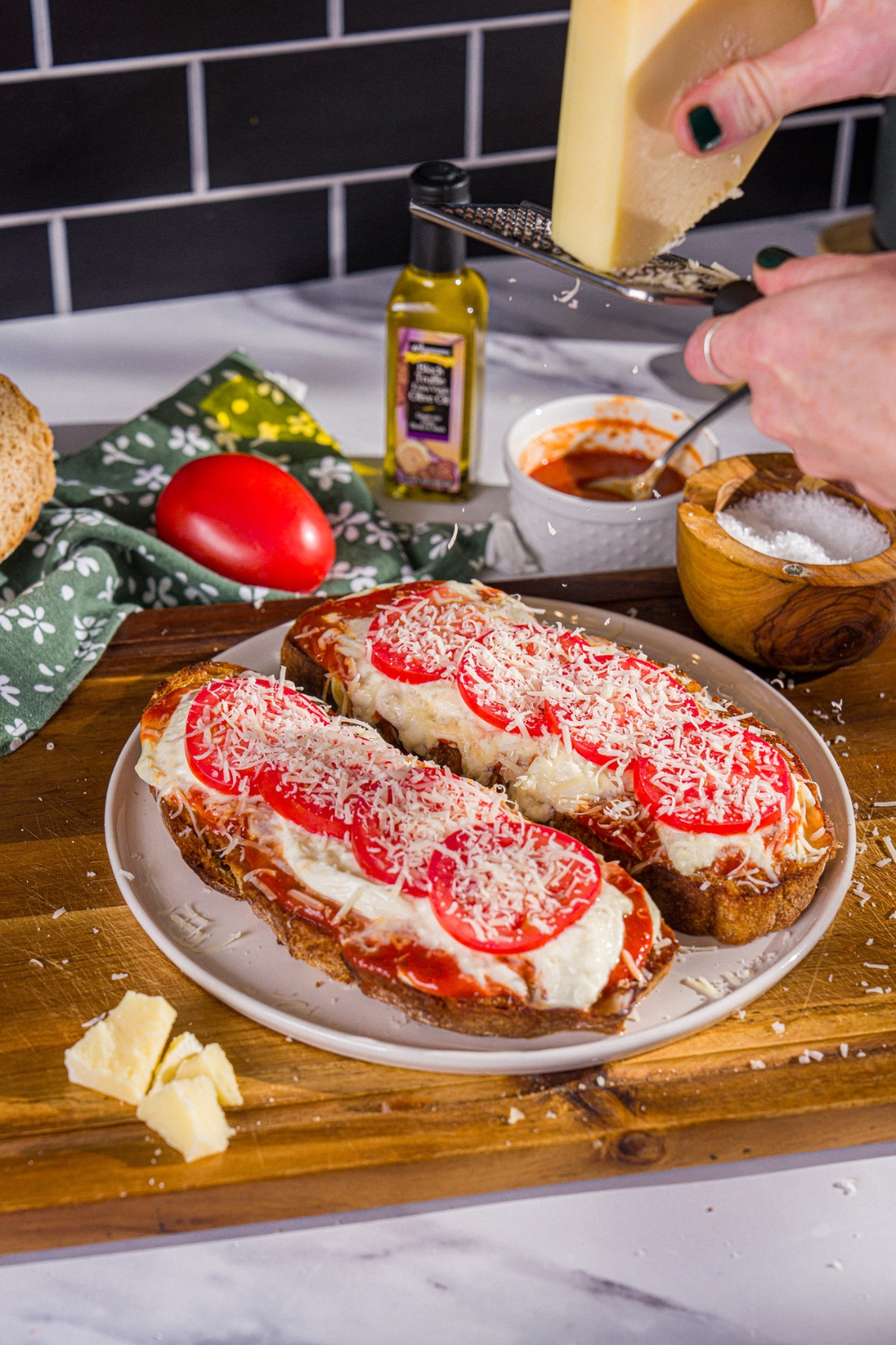 A plate with two slices of Hailey Bieber pizza toast with parmesan cheese being grated over the plate. The plate is on a wooden board with a bowl of salt, marinara sauce, and tomato.