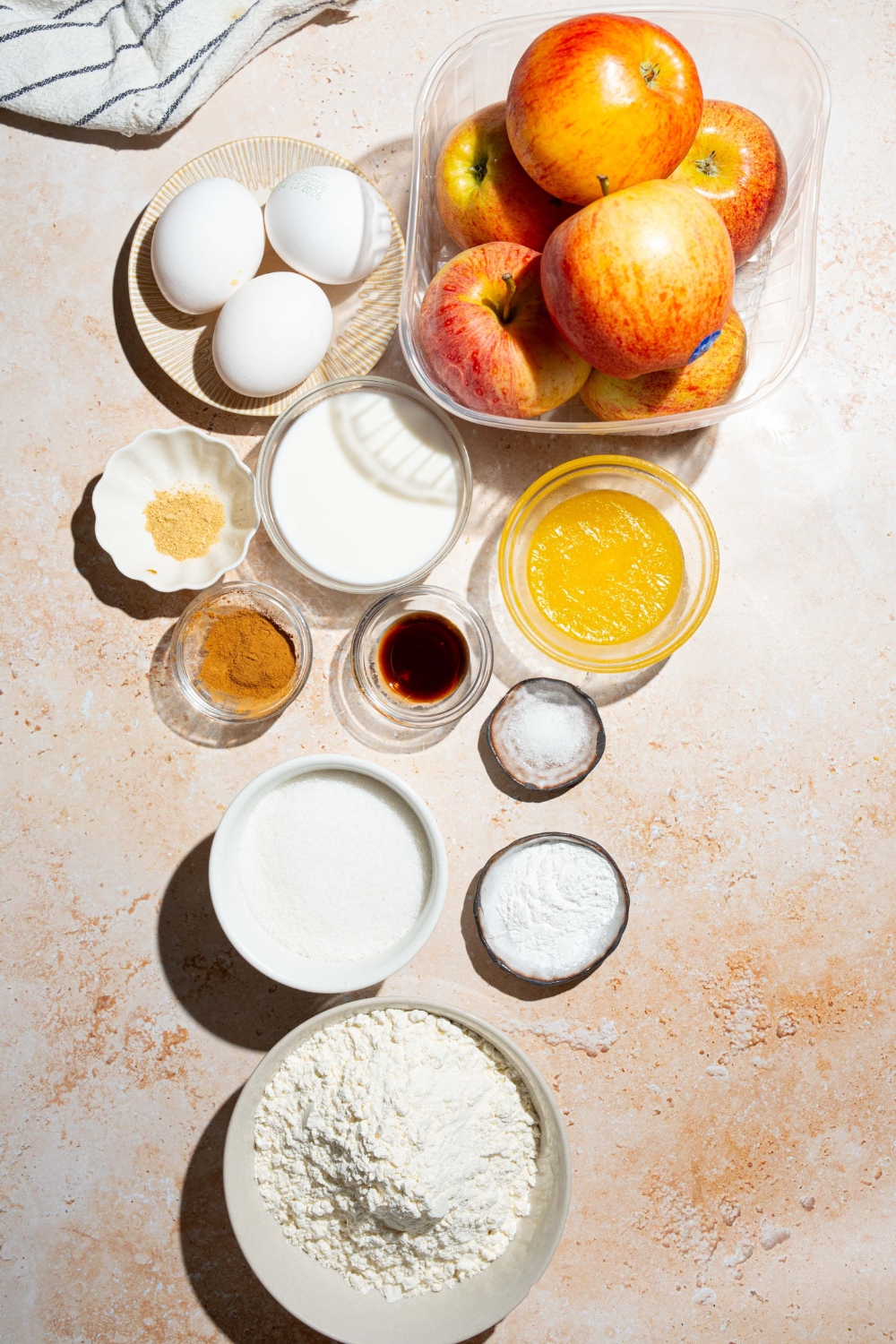 A tan counter with ingredients to make invisible apple cake including apples, flour, vanilla, eggs, milk, sugar, butter, and spices.