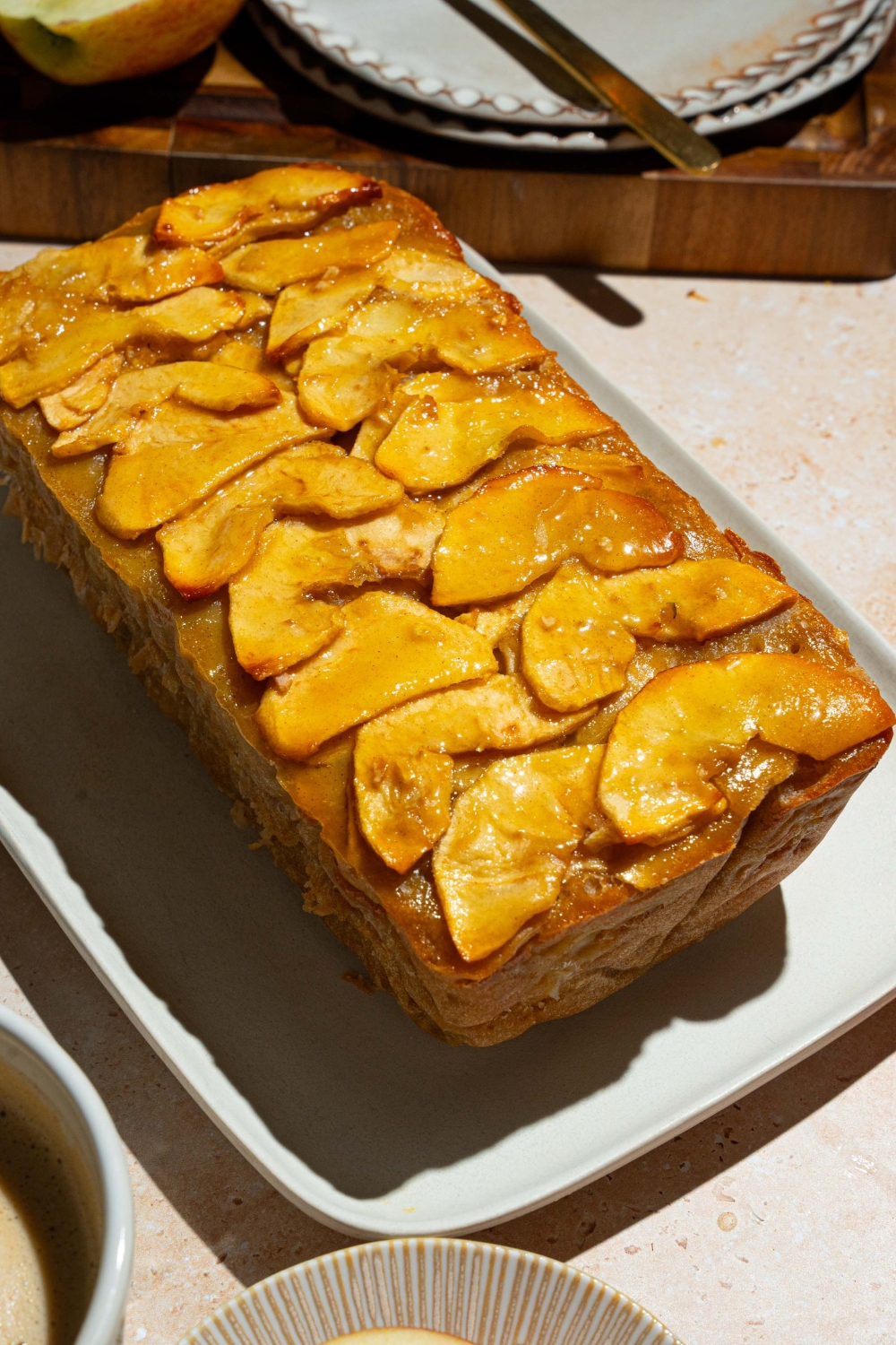 A white platter with a whole invisible apple cake. The platter is on a tan counter with a cutting board and plates.