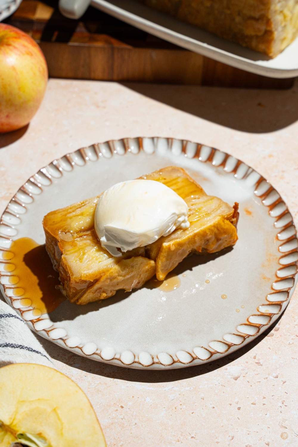 A white plate with a slice of invisible apple cake topped with vanilla ice cream. The plate is on a tan counter with a platter of cake and an apple.