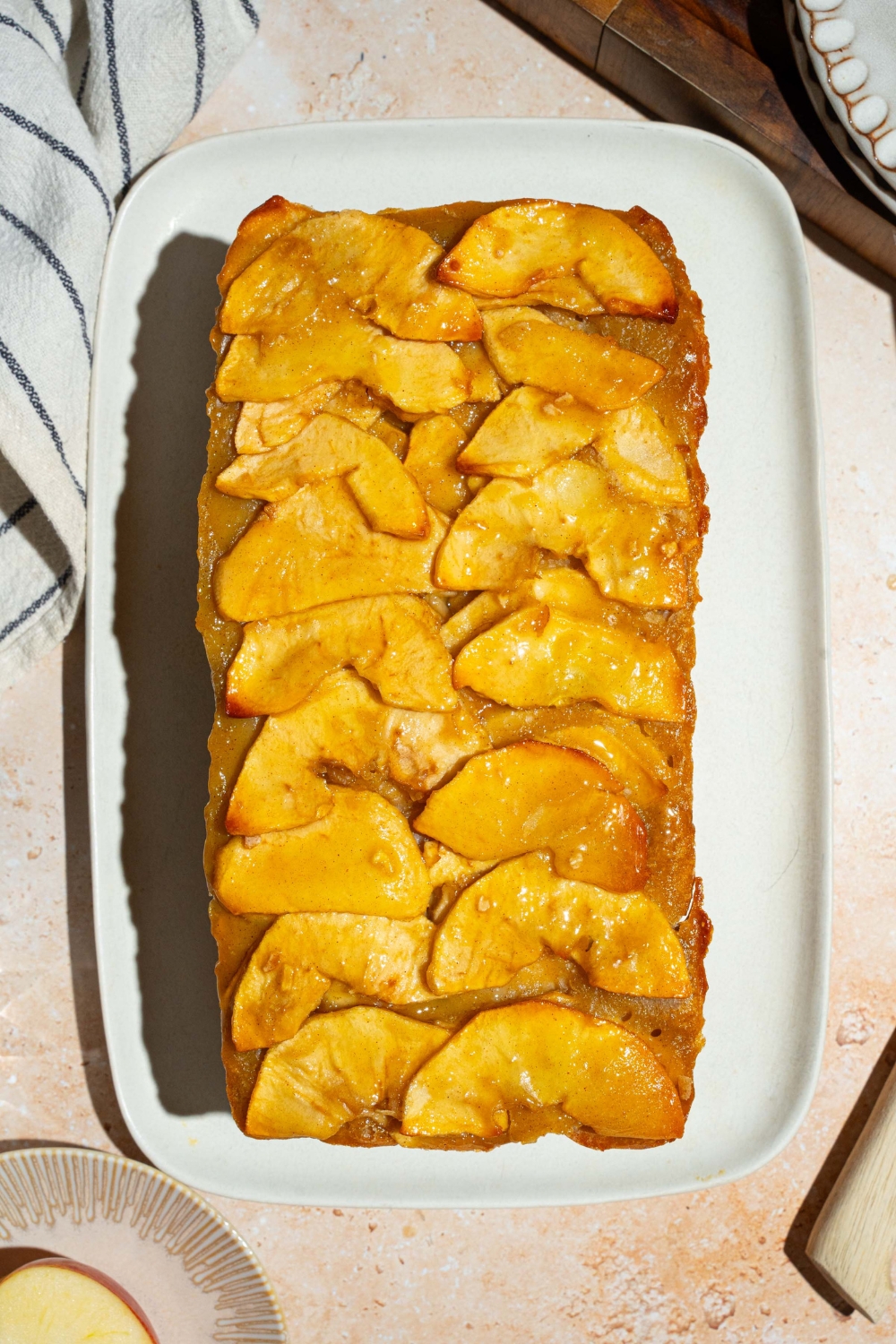 A white platter with a whole invisible apple cake. The platter is on a tan counter with a cutting board and plates.