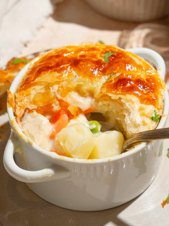 A KFC chicken pot pie in a white ramekin with a golden flaky crust and part of the front of the crust missing showing potatoes, peas, and carrots in a creamy filling.