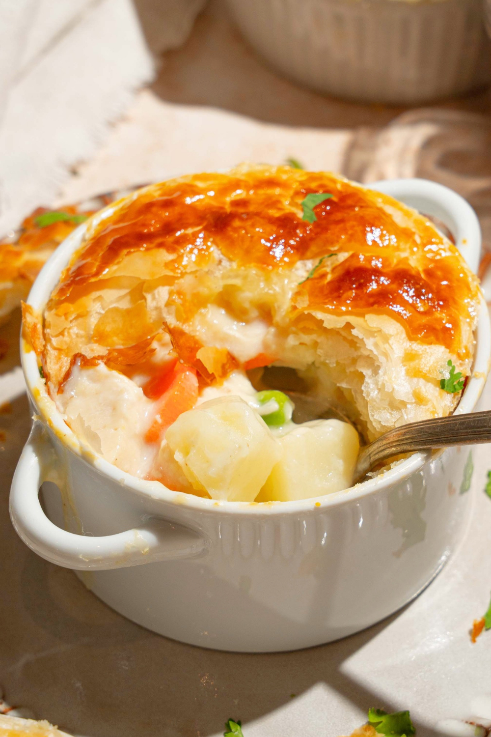 A KFC chicken pot pie in a white ramekin with a golden flaky crust and part of the front of the crust missing showing potatoes, peas, and carrots in a creamy filling.
