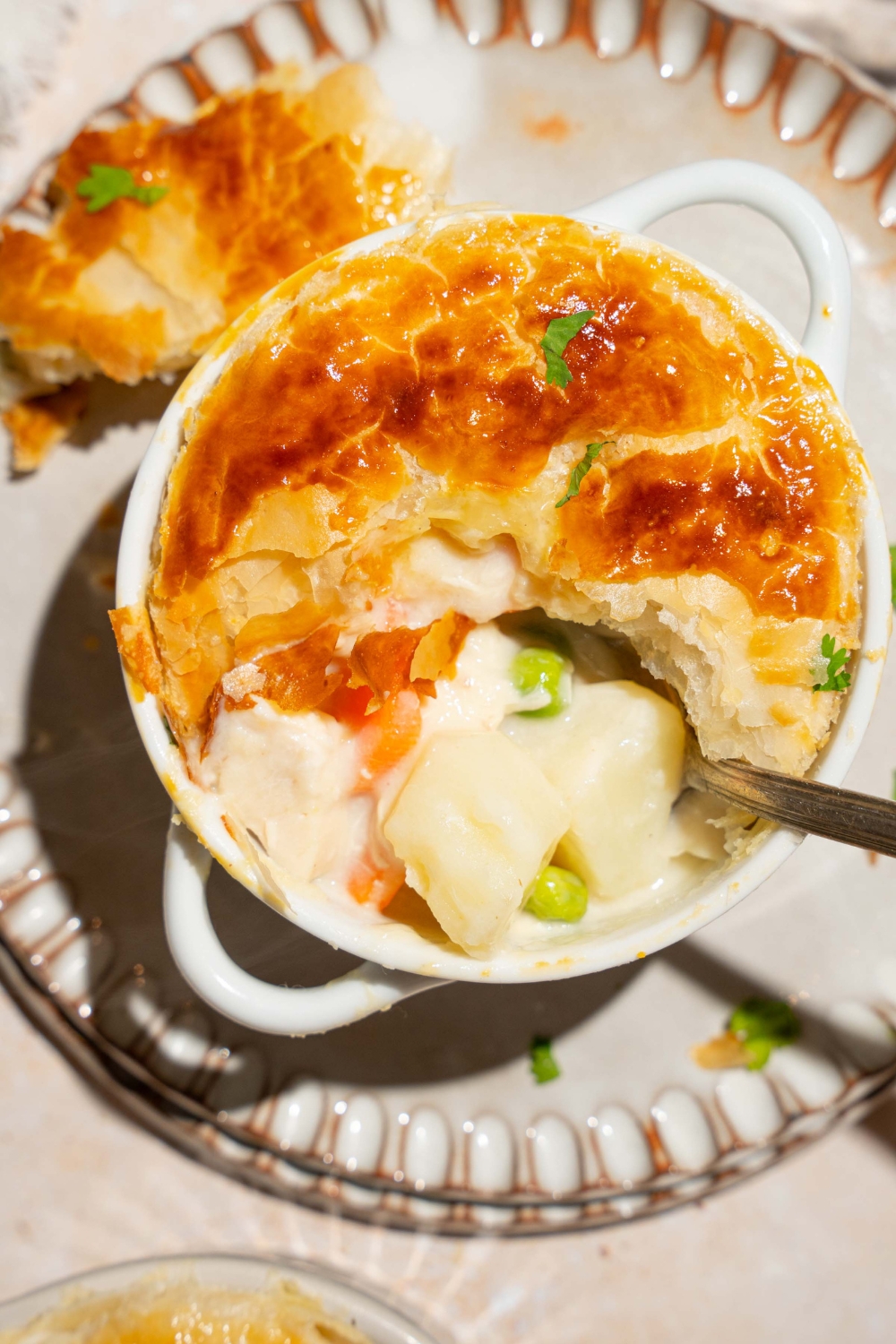 A small pie tin with baked KFC pot pie topped with puff pastry and garnished with fresh parsley. A spoon is taking a bite from the pot pie. The tin is on plate.