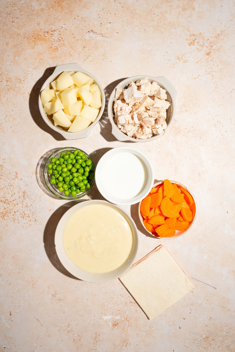 A tan counter with ingredients to make KFC pot pie including puff pastry sheets, shredded chicken, carrots, peas, milk, potatoes, and cream of chicken soup.
